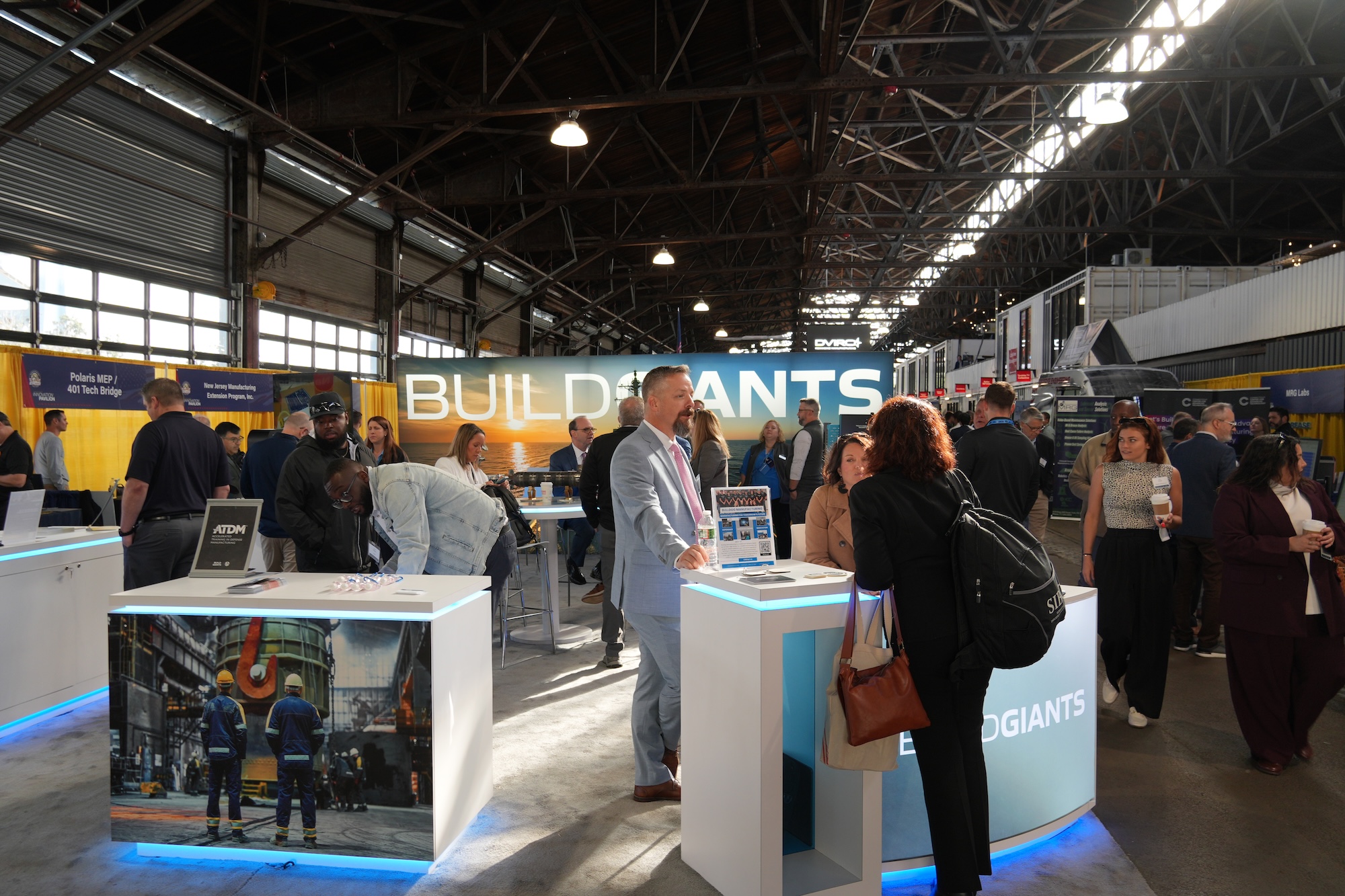 People interact at an indoor exhibition booth with "BUILD GIANTS" signage and digital displays, under industrial-style lighting.