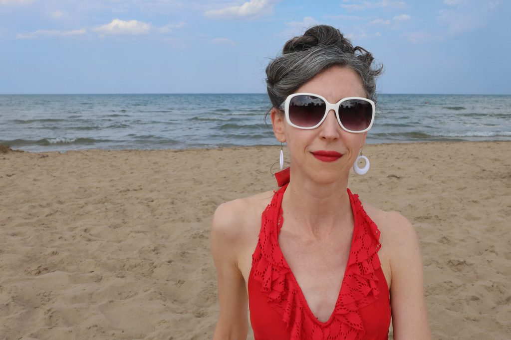 A woman with gray and black hair, wearing white sunglasses, red lipstick, white earrings, and a red halter dress, stands on a sandy beach with the ocean and blue sky in the background.