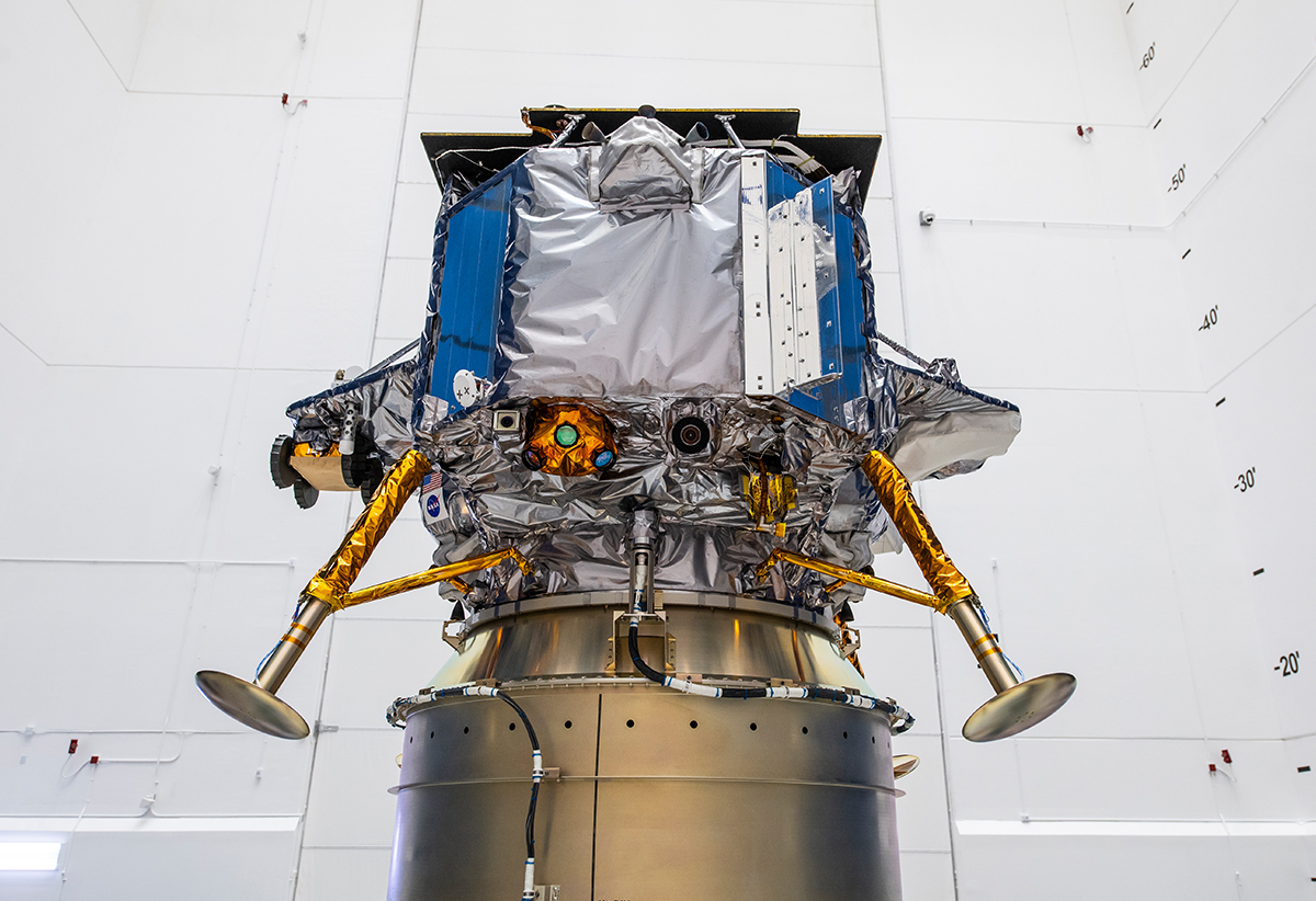 A spacecraft with metallic components, blue panels, and gold-colored landing legs stands in a white, industrial testing facility.