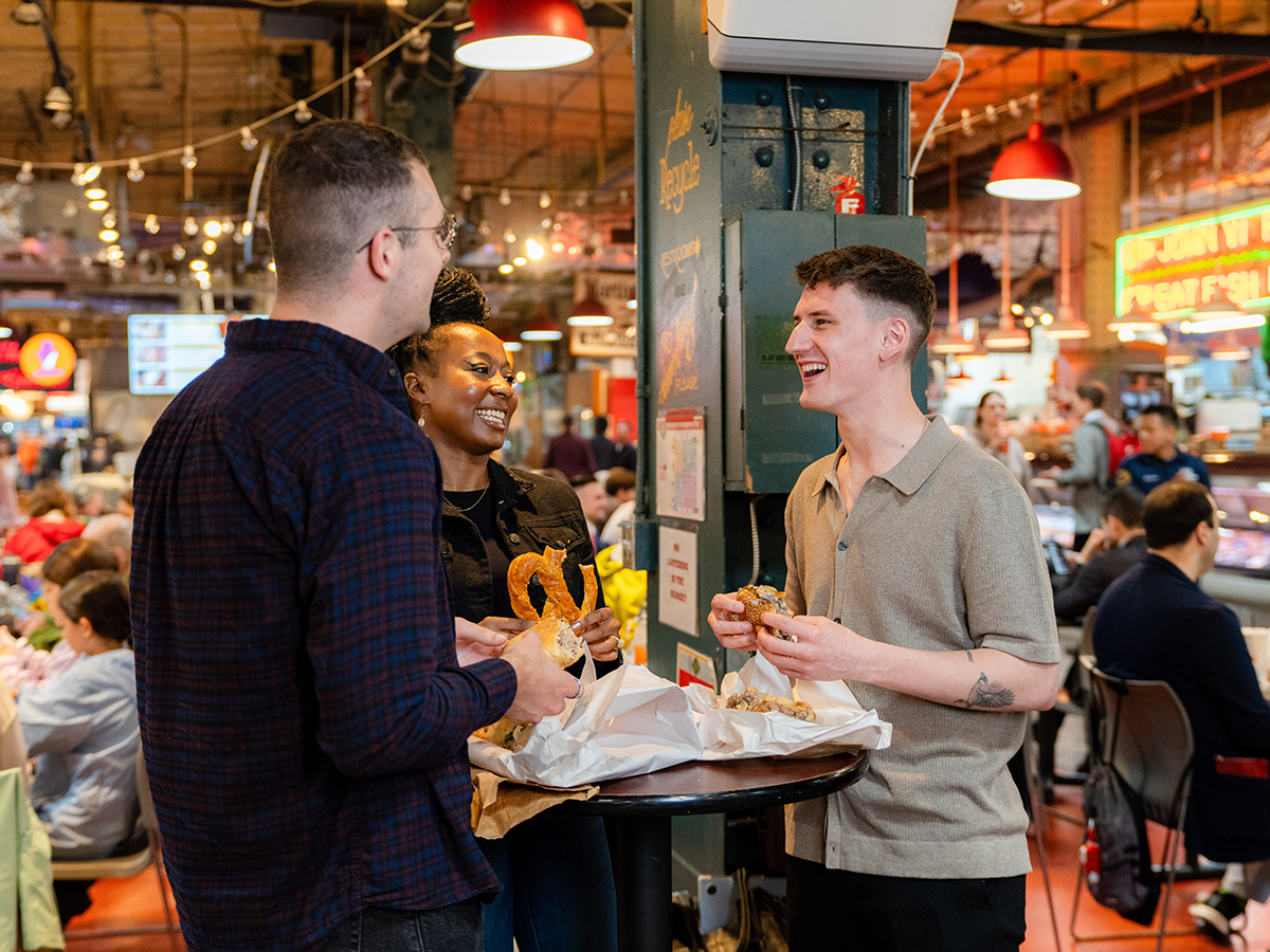 Three people stand around a high top table while eating cheesesteaks and soft pretzels at Reading Terminal Market in Philadelphia.