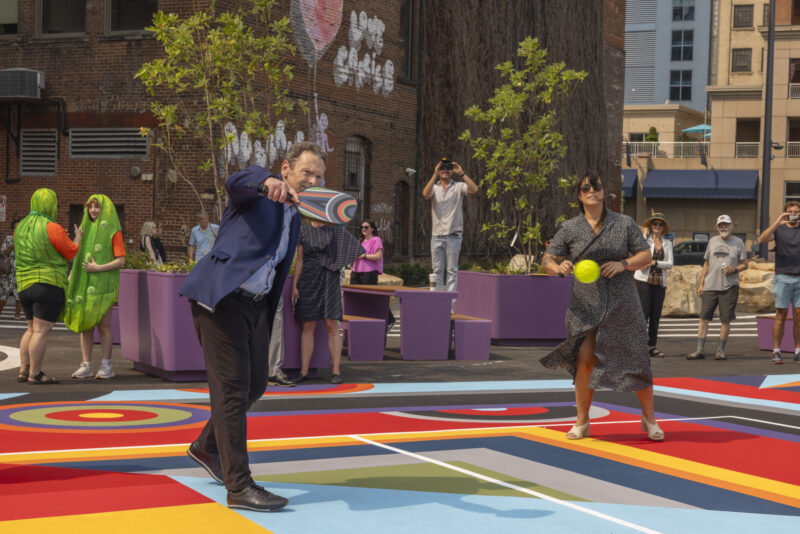 A man in a suit throws a ring while a woman stands beside him holding a ball, in a colorful outdoor urban area with onlookers and people in costumes—a lively scene reminiscent of Sharmistha Ray’s Geometry of Play.