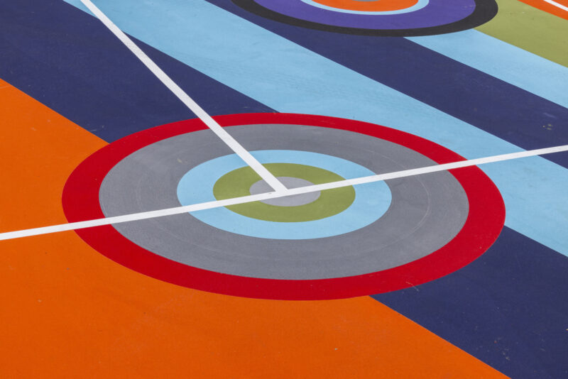 A colorful painted court inspired by Sharmistha Ray Geometry of Play, featuring concentric circles, diagonal stripes in blue tones, and intersecting white lines on an orange background.