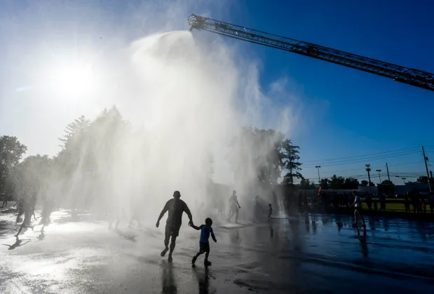 Silhouetted against the hot summer sun, Derek Colonello and son Nathan, 3, run through the cool spray of water Thursday, July 2, 2026, during a free community cool off event featuring Coopersburg Fire Rescue's ladder truck spraying water in Coopersburg. (April Gamiz/The Morning Call)