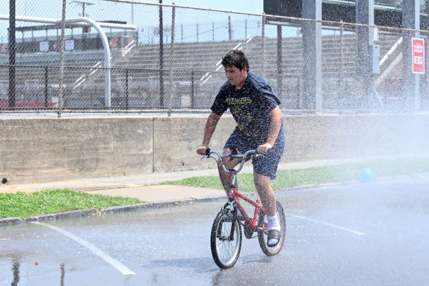 Justin Bahrt, 14, of Easton rides his bicycle through water flowing from an open fire hydrant Wednesday, July 15, 2026, between Cottingham Stadium and Paxinosa Elementary School in Easton. Eight of the city's hydrants will be open again from noon to 6 p.m. Thursday to combat soaring temperatures. (Jon Chiu/The Morning Call)