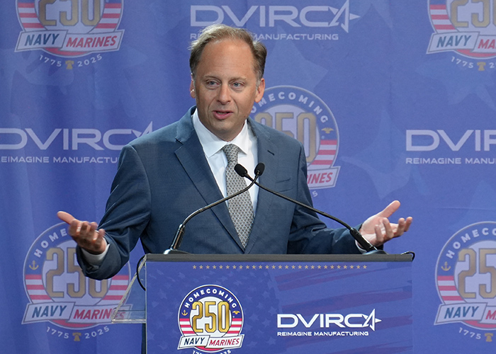 A man in a suit speaks at a podium with a microphone, in front of a Navy and Marines 250th anniversary event backdrop.