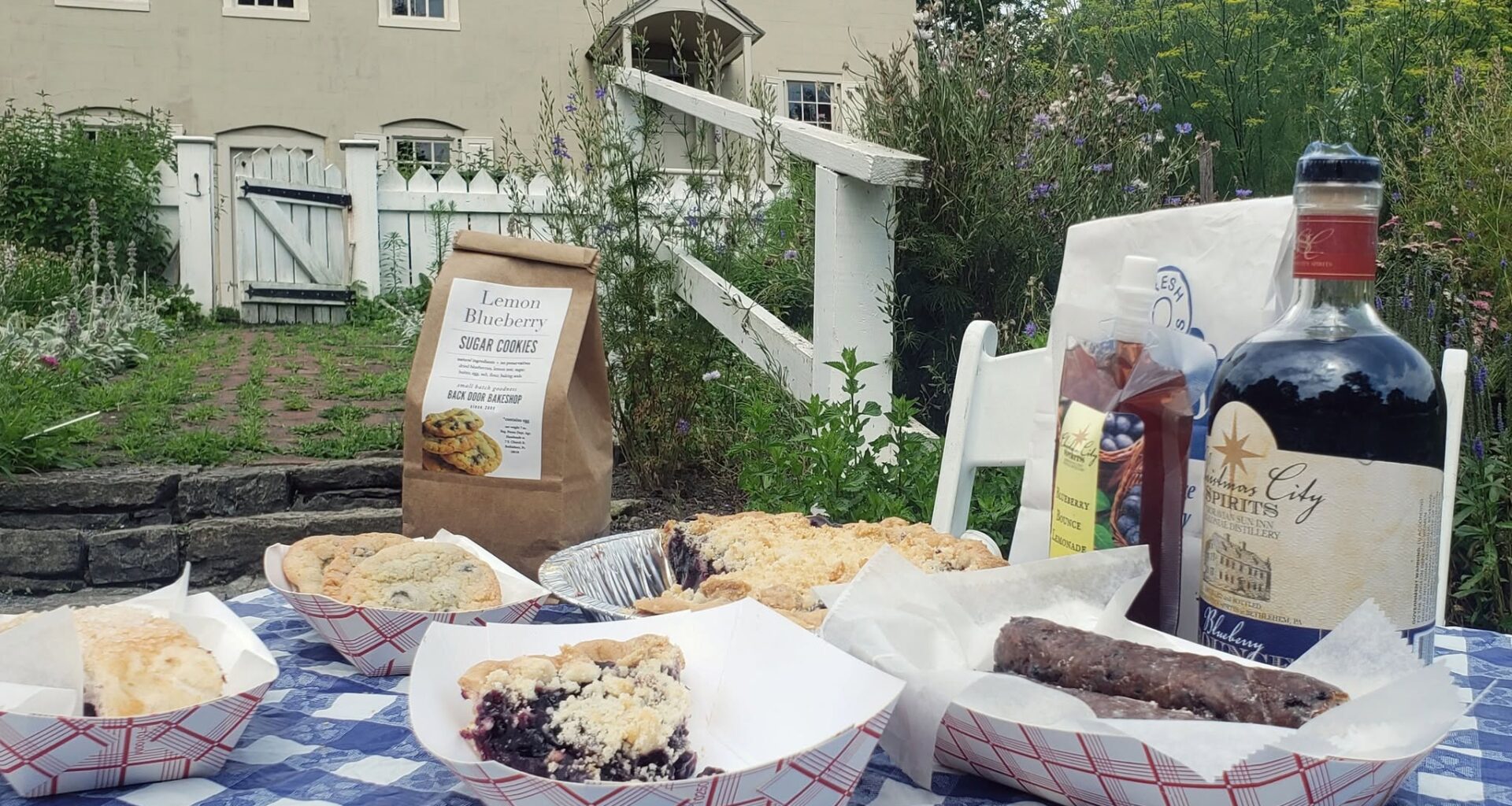 Blueberry desserts and drinks sit on a checkered tablecloth outside a historic farmhouse.