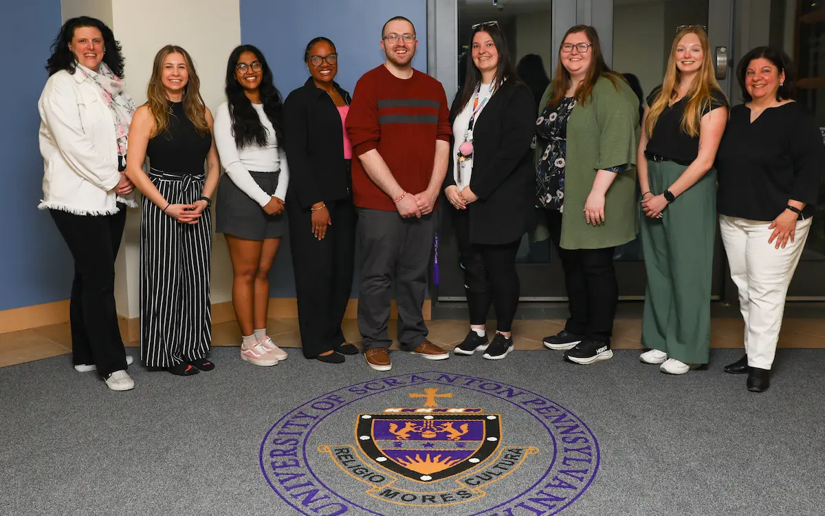 Nine people pose together indoors, standing on carpet with the University of Scranton seal visible on the floor.