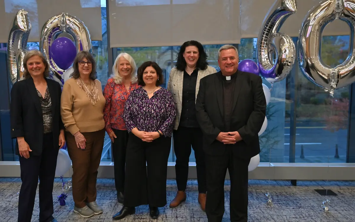 Group of six adults smiling at an indoor celebration, standing in front of large silver “60” balloons and purple-and-white decorations.