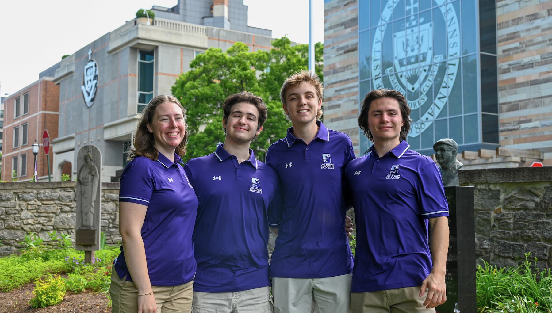 Four students in matching purple polo shirts pose outdoors in front of campus buildings and a large university seal on a glass facade.