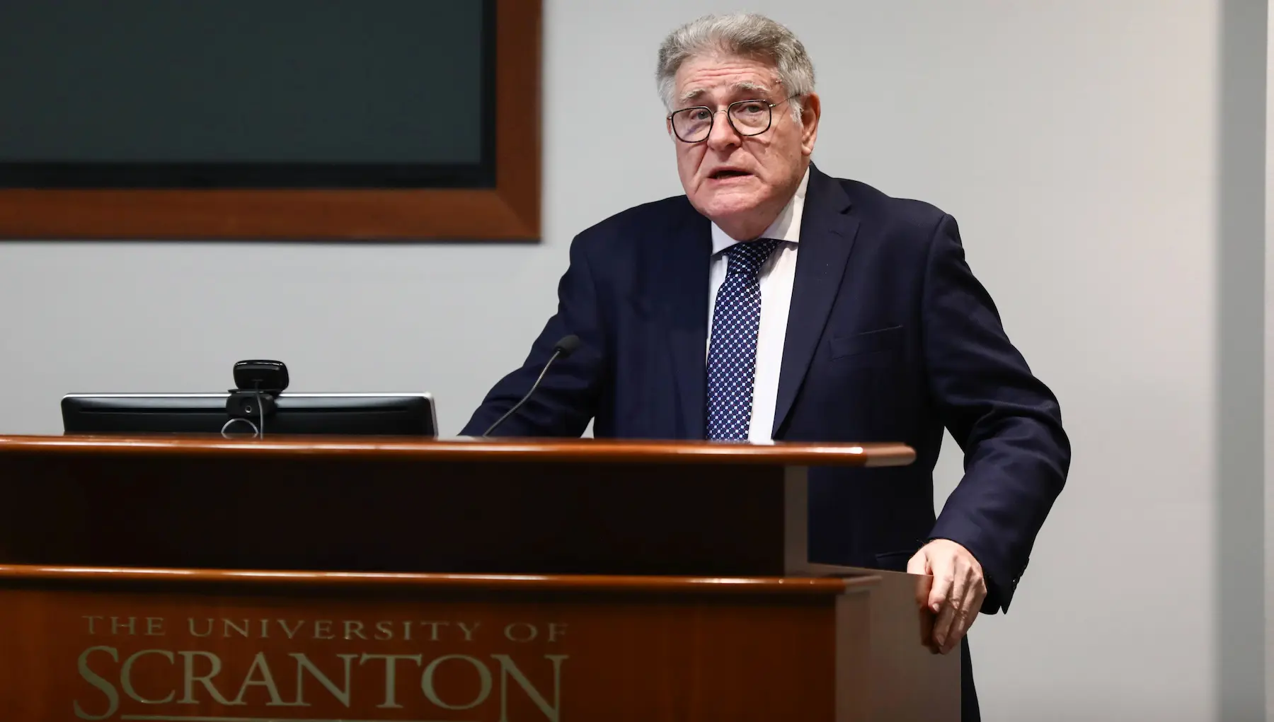 Individual in a suit and glasses speaks at a podium labeled "The University of Scranton" in a lecture hall.
