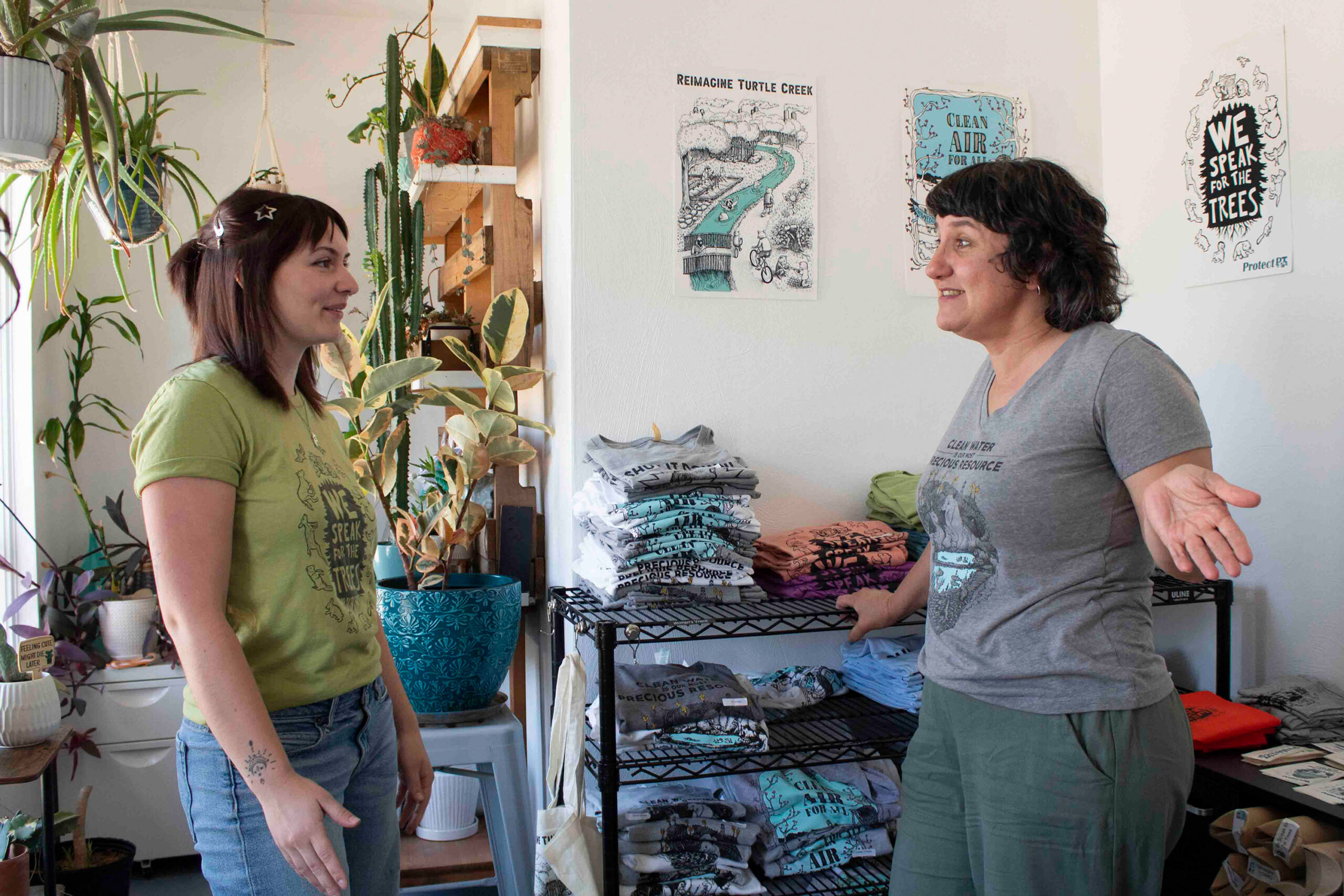 Two women stand and talk in a small shop, surrounded by folded t-shirts, plants, and posters on the wall.