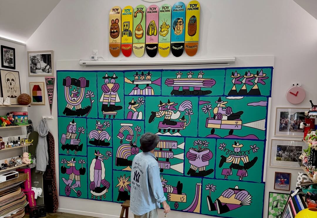 An artist stands barefoot in a studio looking at a large green mural depicting cartoon characters.