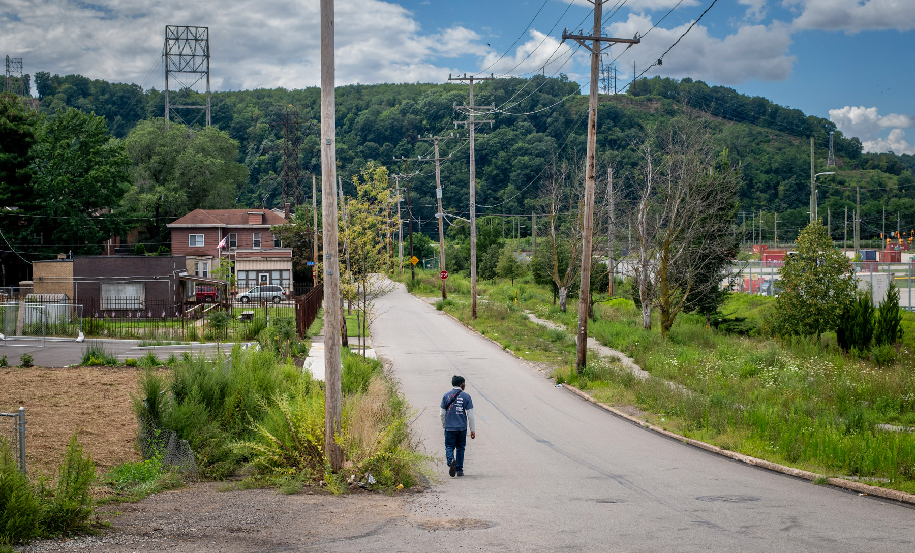 A person walks alone down a quiet, empty street lined with overgrown grass and utility poles, with houses and hills in the background under a partly cloudy sky.