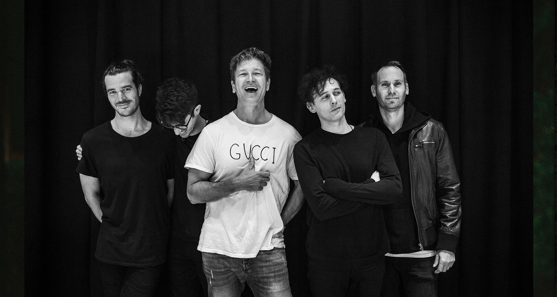 Five men stand together smiling and laughing against a black curtain in a black-and-white photo.