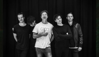 Five men stand together smiling and laughing against a black curtain in a black-and-white photo.