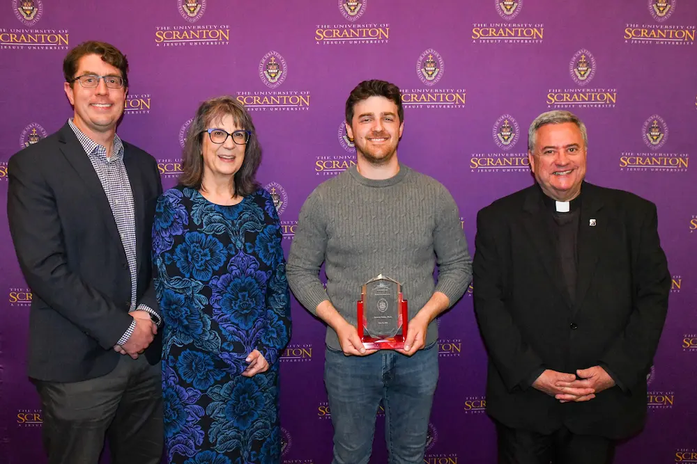 Four people pose in front of a University of Scranton step-and-repeat backdrop, with one person holding a glass award plaque.