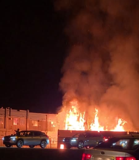 Several cars are parked in front of a building at night. Flames and smoke are rising from the building.