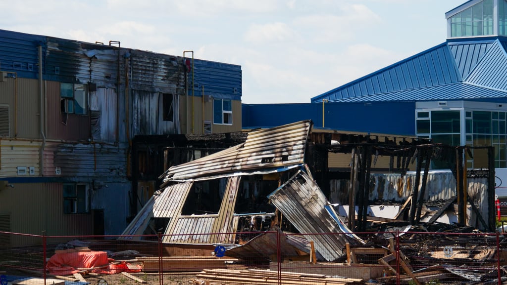 Walls of a portable classroom are charred and falling over in front of a school building.