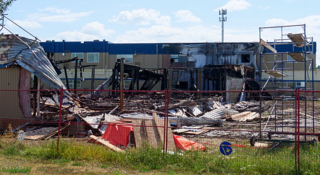 Behind a fence, charred poles are left standing in front of a burned school exterior surrounded by pieces of wall and metal.