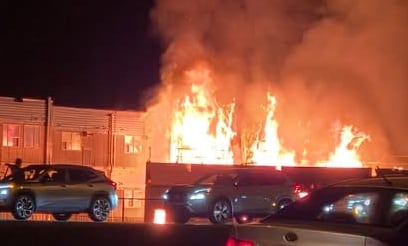 Several cars are parked in front of a building at night. Flames and smoke are rising from the building.