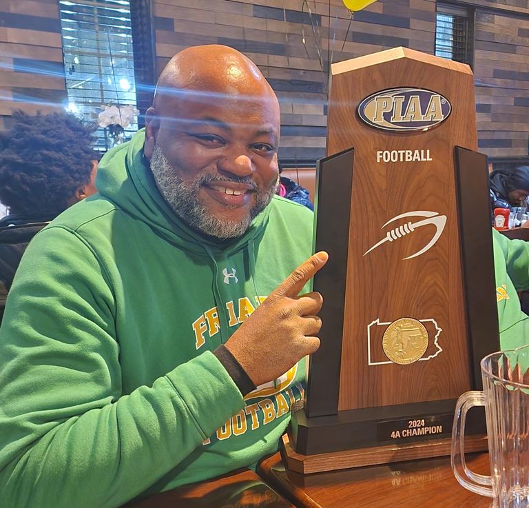 Man in a green hoodie smiling and pointing at a PIAA Football 4A Champion trophy in a restaurant setting.