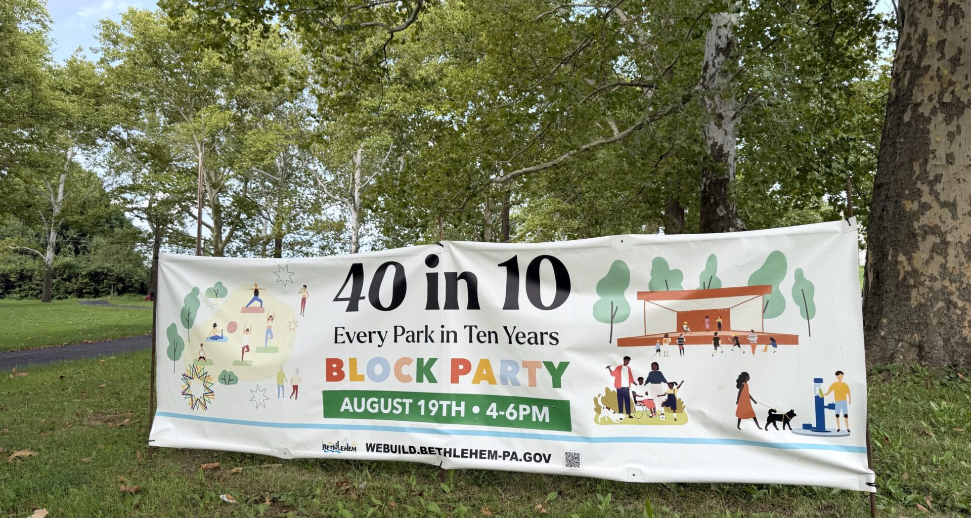 A large white promotional banner stands on a grassy lawn surrounded by trees in a park.