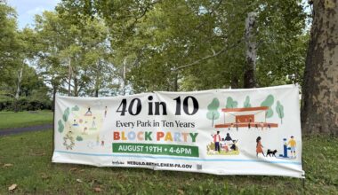 A large white promotional banner stands on a grassy lawn surrounded by trees in a park.