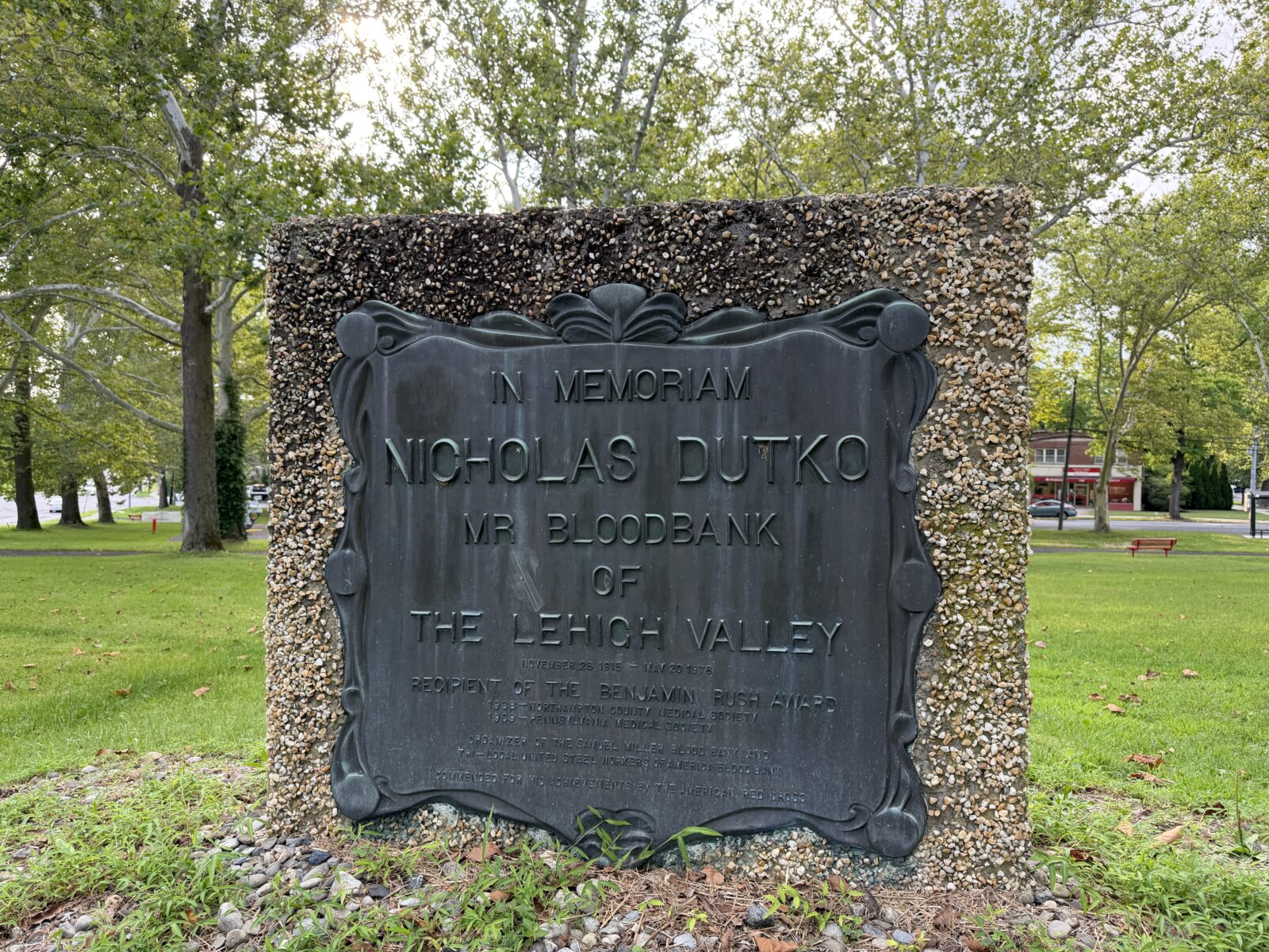 A stone monument holding a bronze plaque honoring Nicholas Dutko in a grassy, tree-lined park.