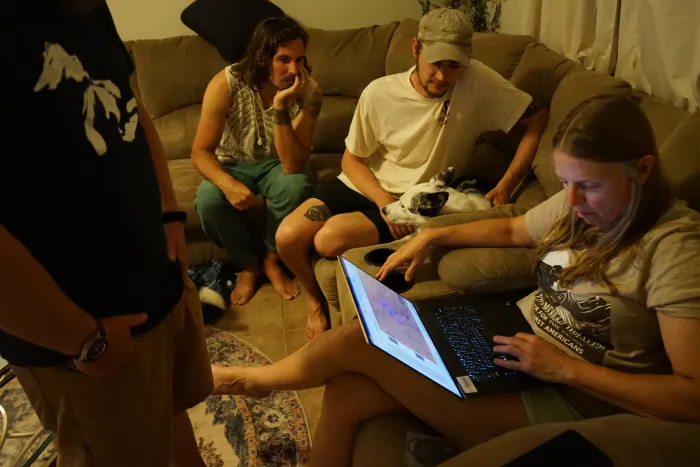 Three people sit on a couch with a dog, looking at a laptop. One person stands in front of them.