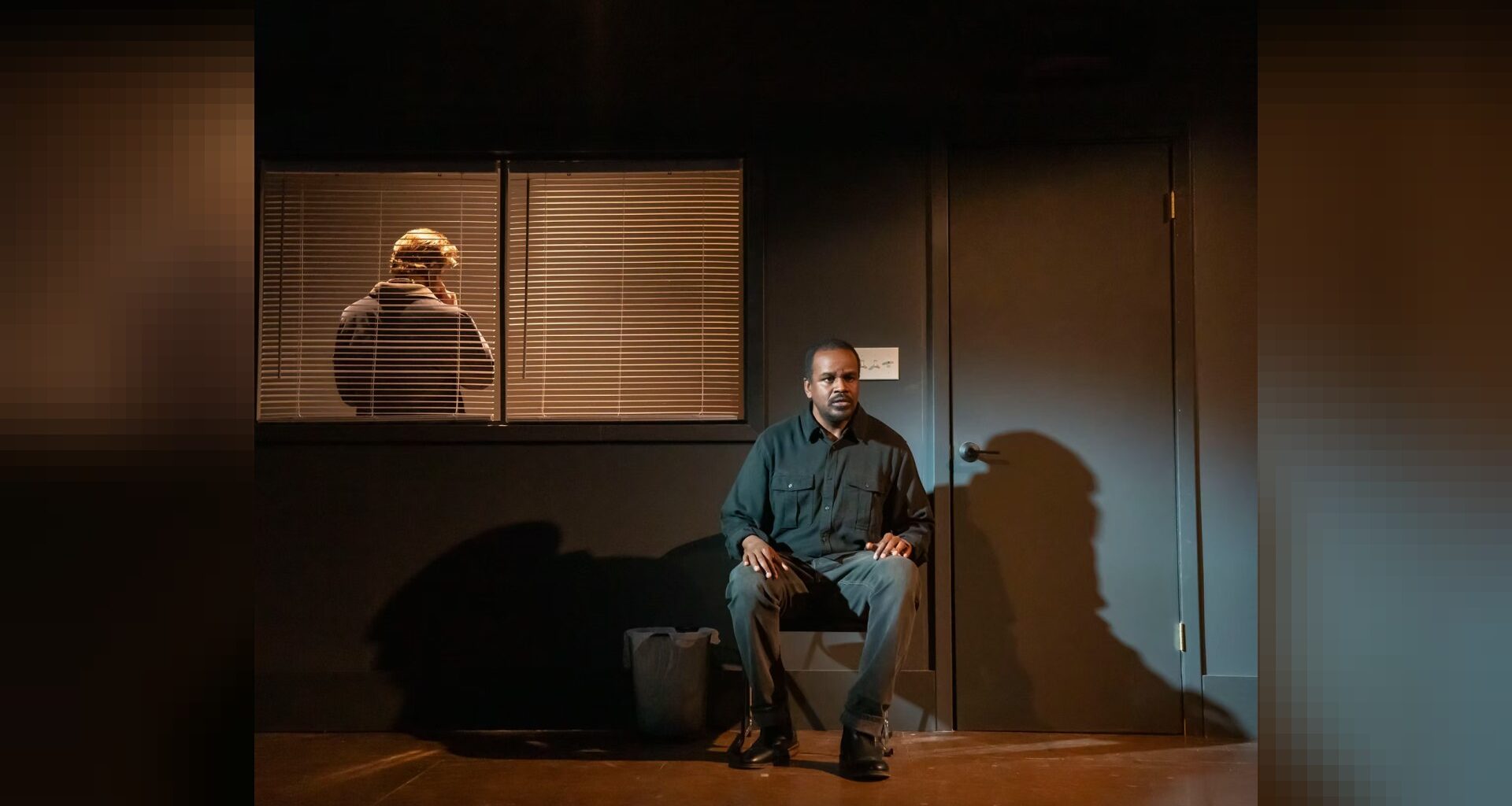 An actor sits on a dark stage next to a closed door while another actor stands behind window blinds.
