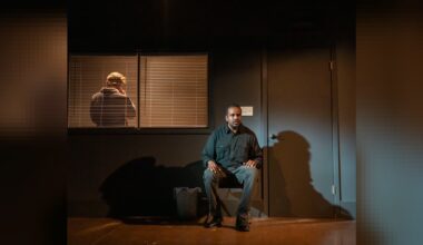 An actor sits on a dark stage next to a closed door while another actor stands behind window blinds.