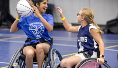 An individual in a wheelchair holds a basketball in the air and smiles. Another person to her right reaches out toward the ball.