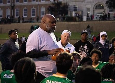 A coach talks to a group of young football players wearing green jerseys on a field, with a large building visible in the background.