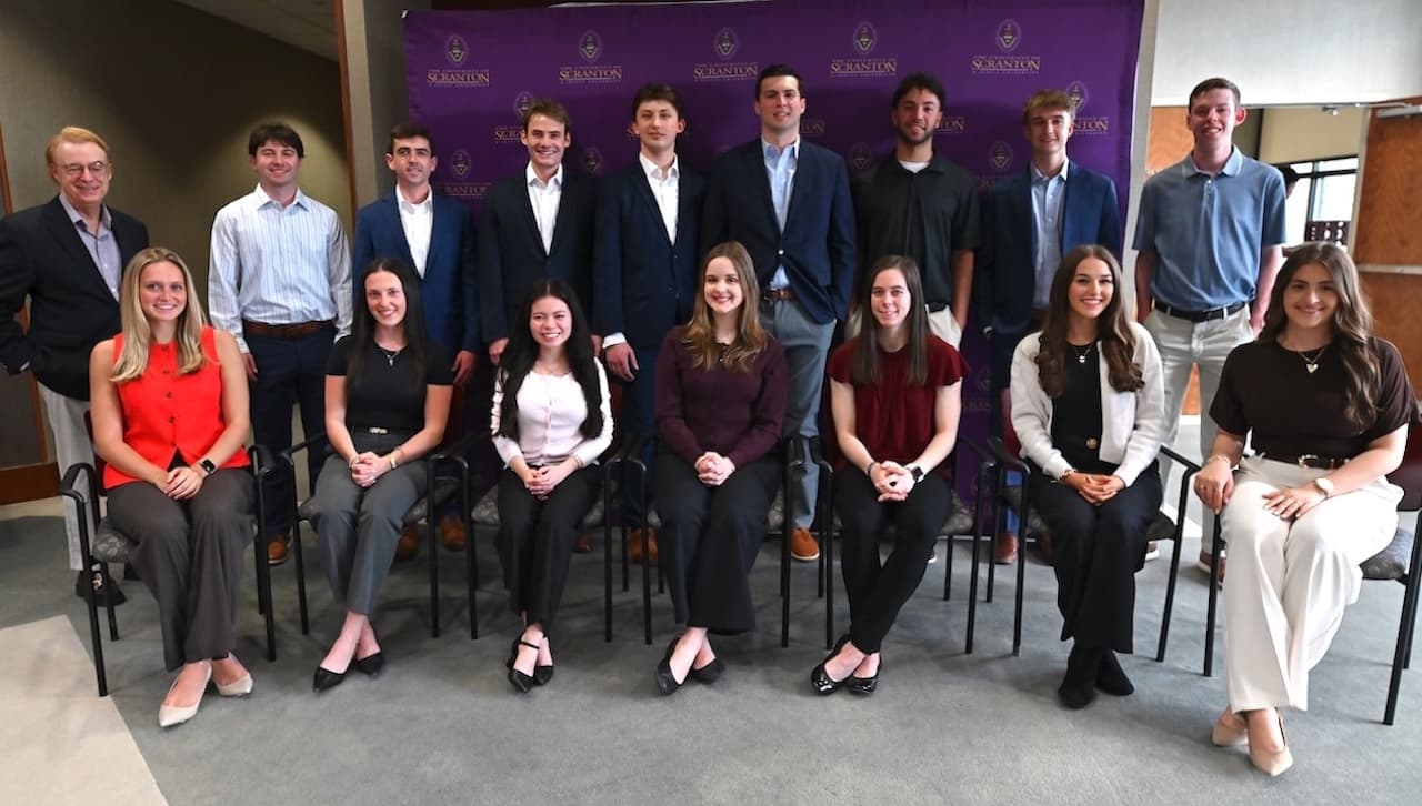 Group of students and faculty posed for a formal photo in front of a University of Scranton backdrop, with some seated in chairs and others standing behind.