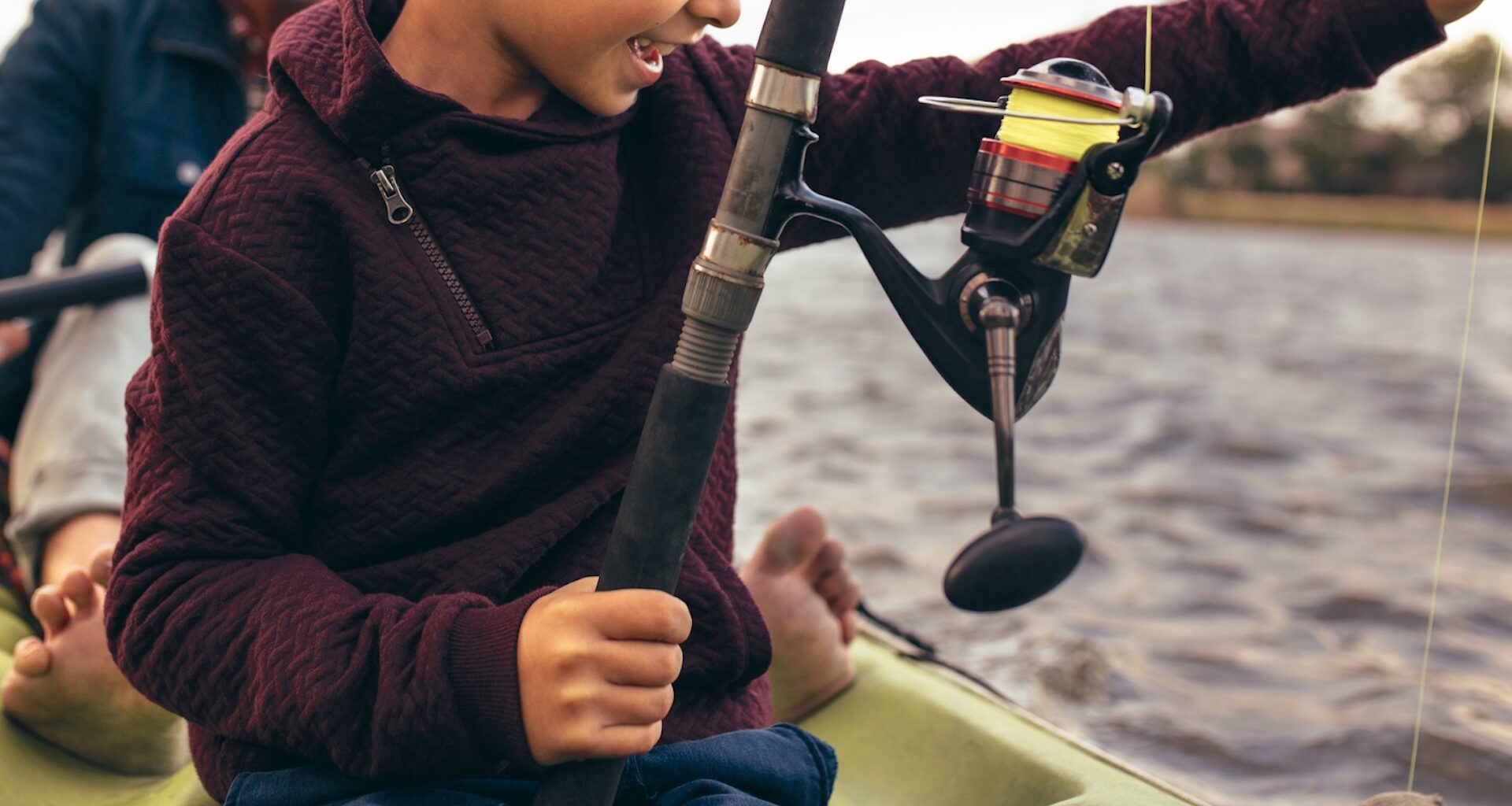 A child holding a fishing rod on a kayak with water in the background.