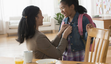Mother buttons daughter’s jacket as she gets ready for school. Daughter is wearing a backpack. A table with breakfast dishes is in front of them.