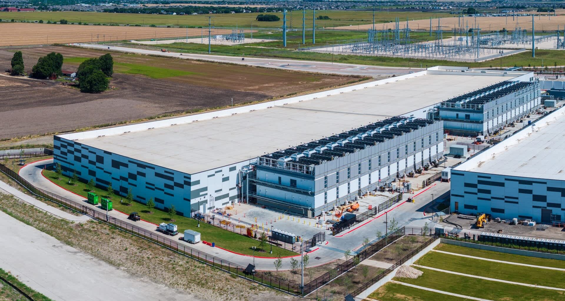 An industrial building with a large flat roof and several loading docks, surrounded by fields and power lines.