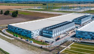An industrial building with a large flat roof and several loading docks, surrounded by fields and power lines.