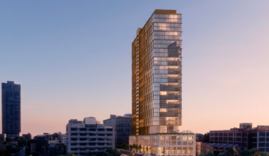 Tall modern skyscraper with glass facade in an urban setting, captured during sunset, with buildings and a road in the foreground.