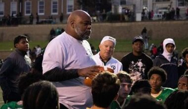 A coach talks to a group of young football players wearing green jerseys on a field, with a large building visible in the background.