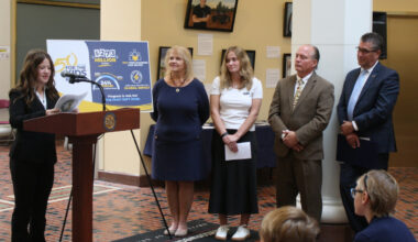 A teenage girl stands at a lectern delivering remarks. Four people stand nearby, looking on, as others – seated in front of her – listen to the comments.