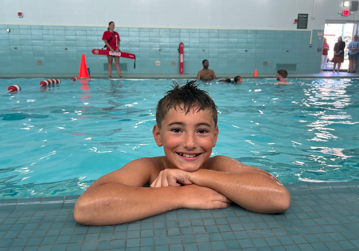 A smiling boy rests at the edge of a YMCA swimming pool after swimming.