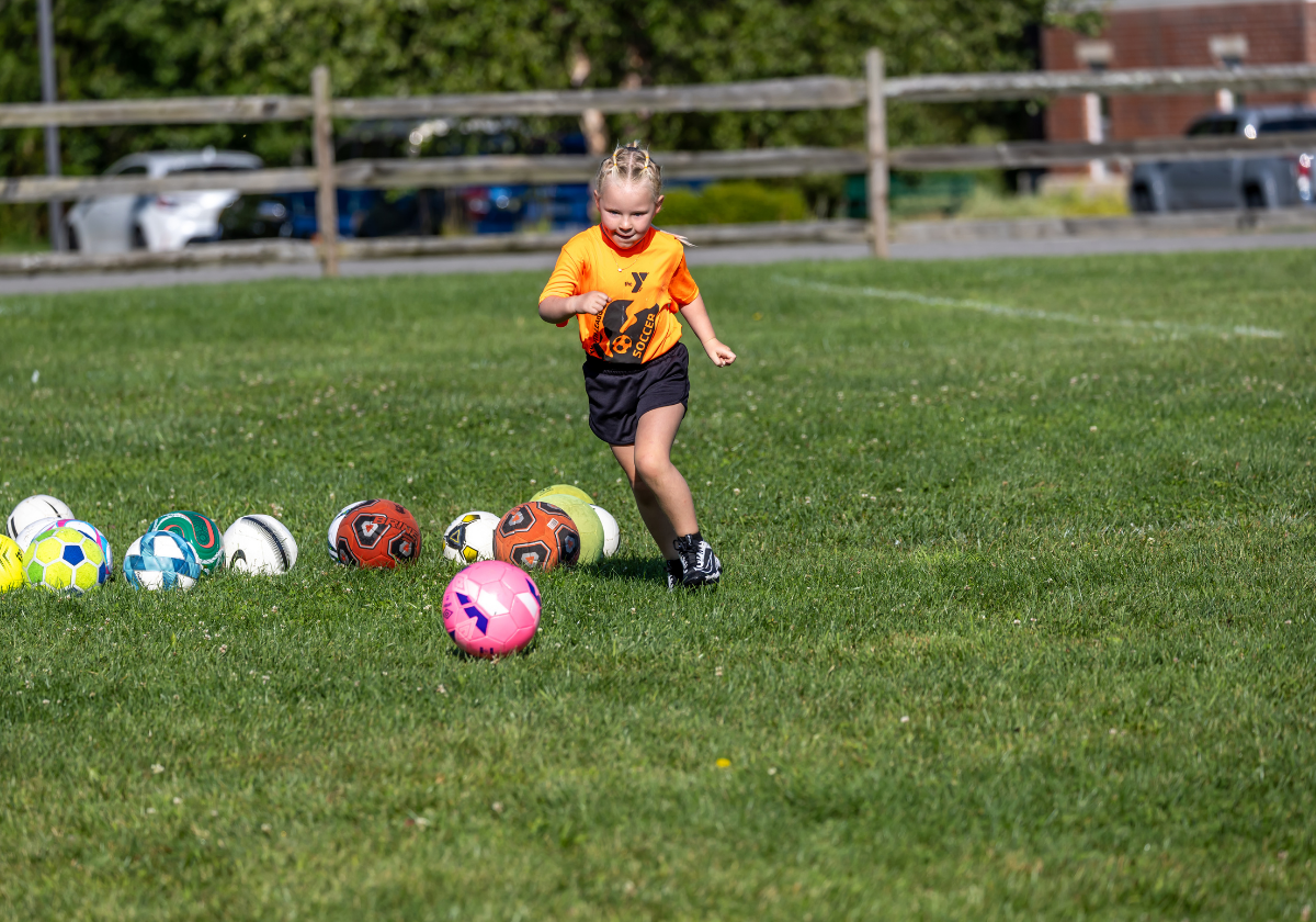 A young child practices soccer on a field during a YMCA youth sports program.