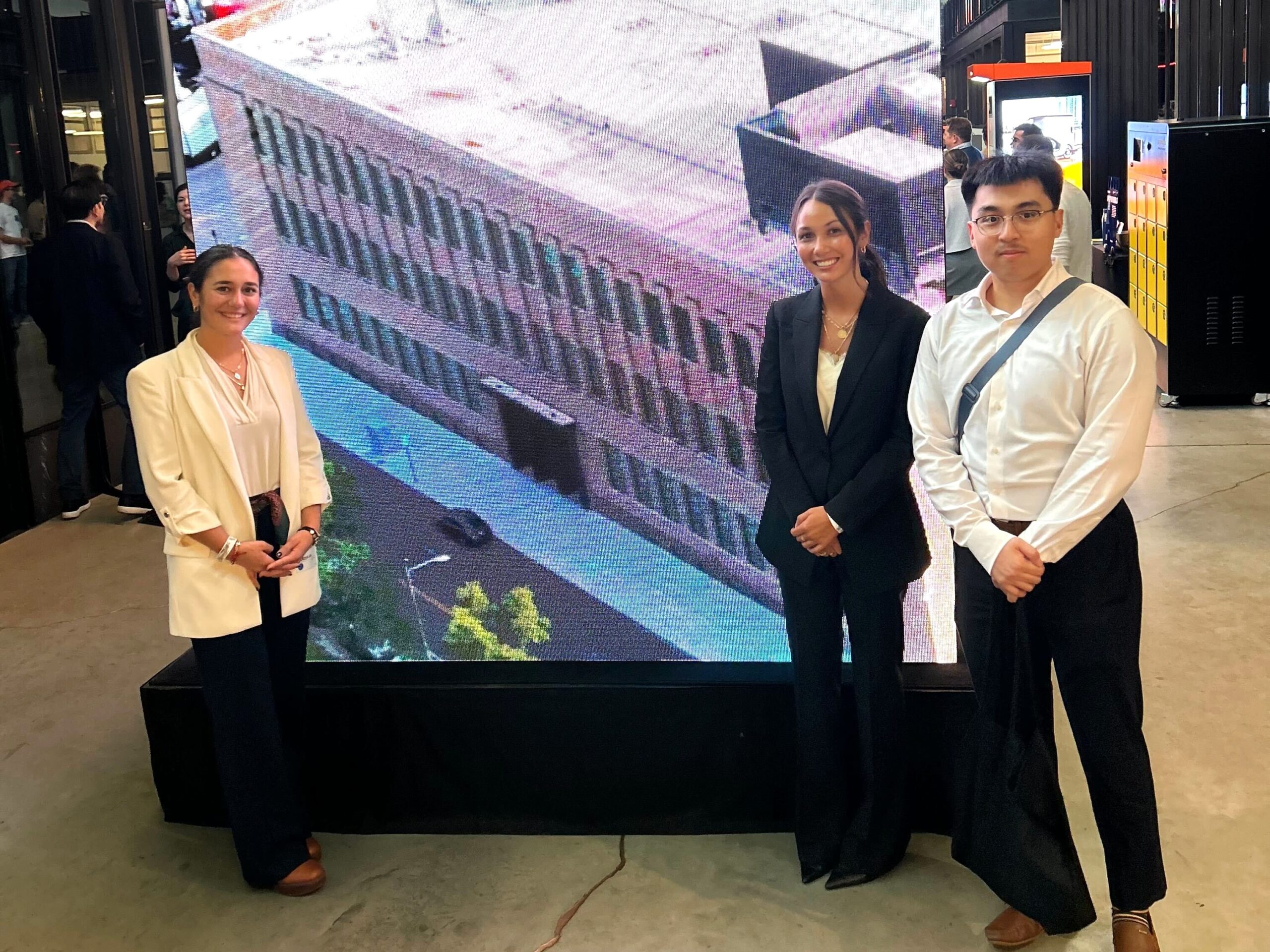 Three people in business attire stand indoors in front of a large digital screen displaying an aerial view of a building.