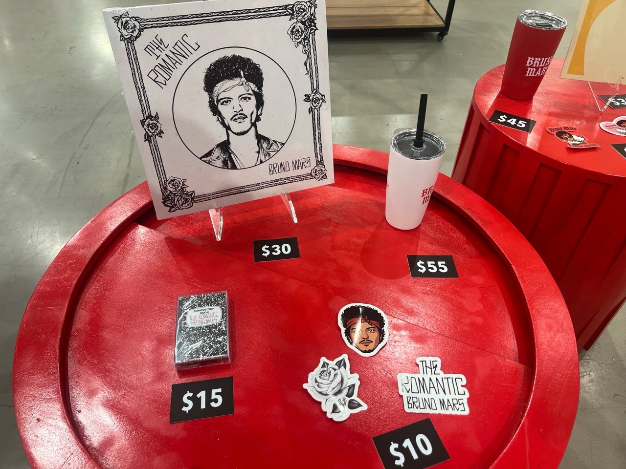 A red table with a number of the smaller and more affordable merch items in the Bruno Mars and Hello Kitty Merch Collaboration pop-up shop. A tumbler, stickers, an LP, and a cassette tape.