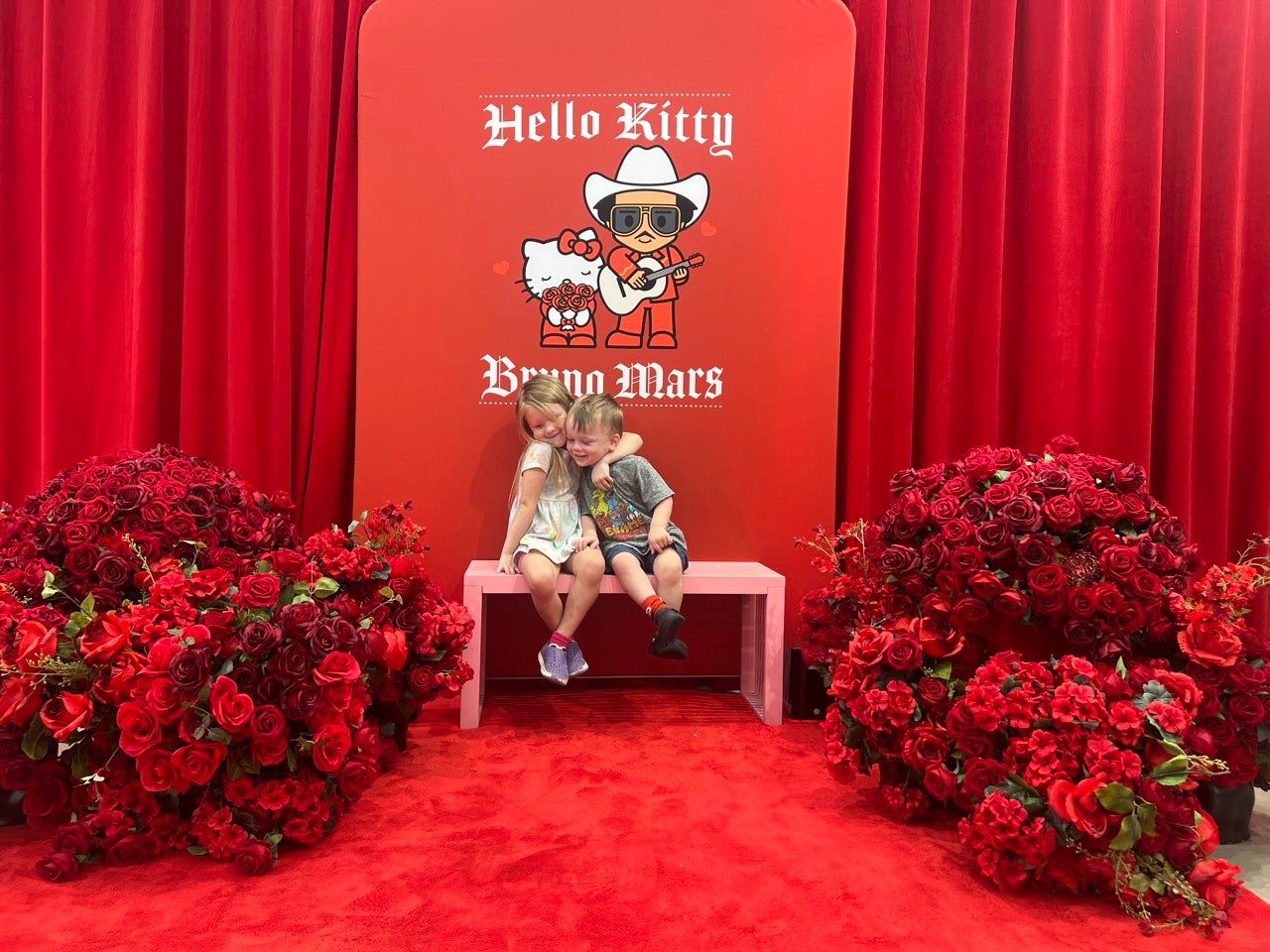 Bruno Mars and Hello Kitty pop up merch store photo op with Kristen's kids sitting on a pink bench surrounded by red flowers and a red backgroung.