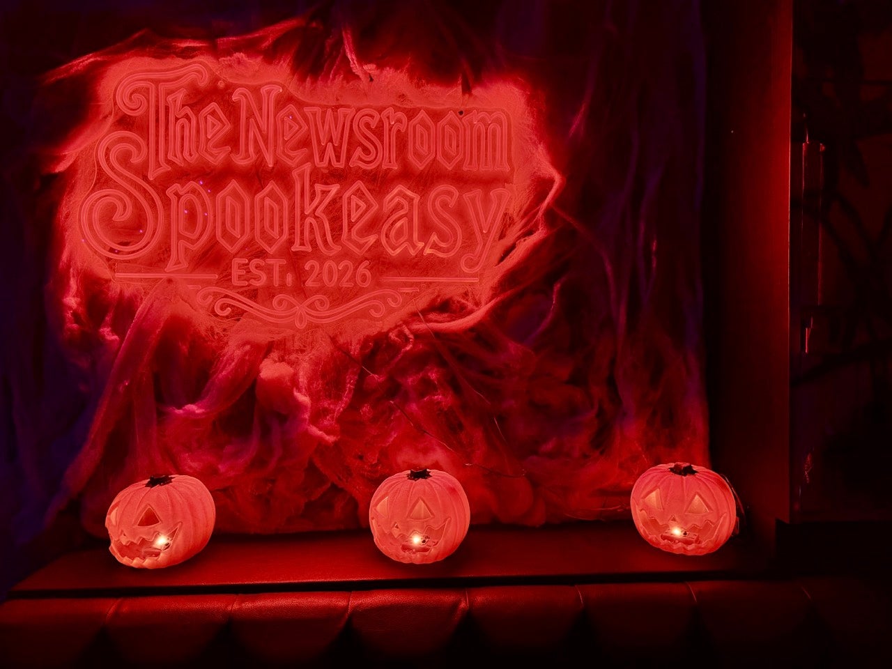 A glowing red sign reads The Newsroom Phialdelphia Speakeasy with 3 glowing pumpkins set beneath it.
