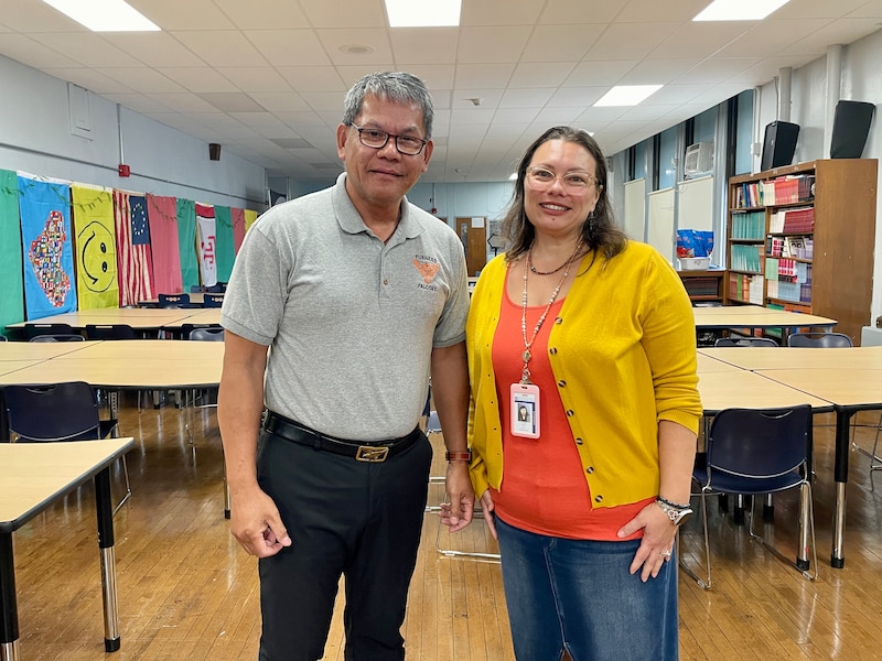 Two adults stand side by side posing for a photograph in a classroom.