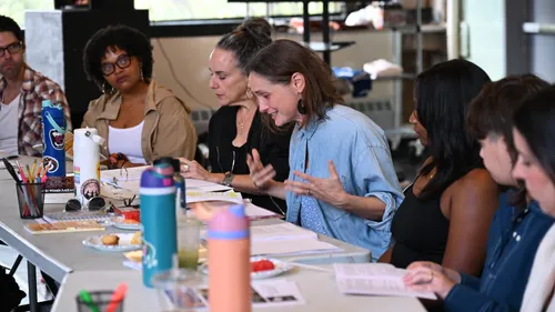 Seven people in casual clothes around a table crowded with scripts, water bottles, and pens focus on a reading.