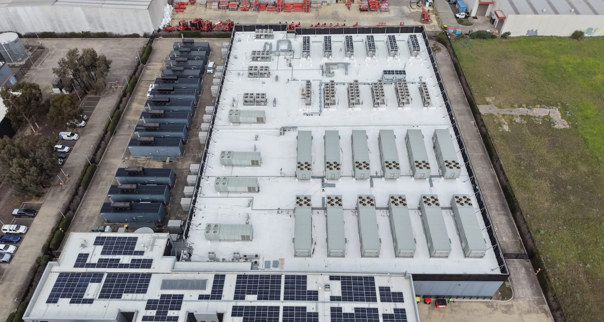 Aerial view of a data center building with rooftop air conditioning units and solar panels, surrounded by vehicles and open space.
